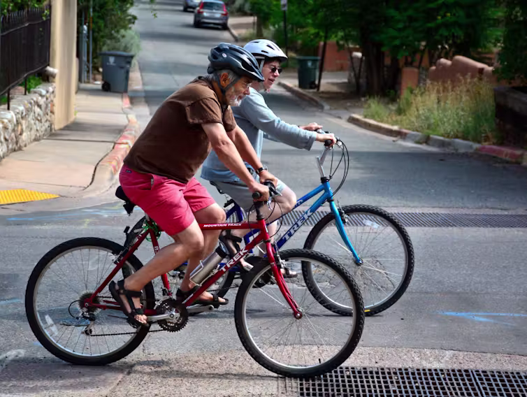 An elderly man and an elderly woman ride their bikes on a street while wearing helmets.