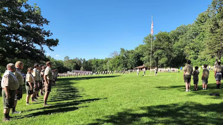 People of all ages in beige uniforms stand in a line in a green field on a sunny day before an American flag on a pole.