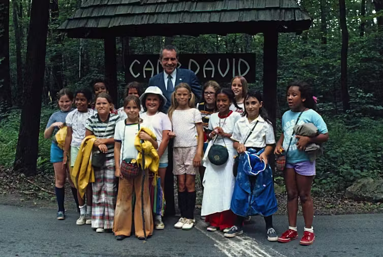 Girls of all races wearing colorful outfits pose with an older man wearing a suit in front of a sign reading 'Camp David.'