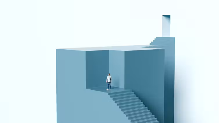 Man standing at the top of a staircase blocked by a solid wall looking toward a door located above him.