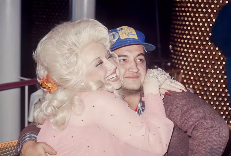 Young woman with blonde hair smiles while hugging a young man wearing a blue baseball cap.